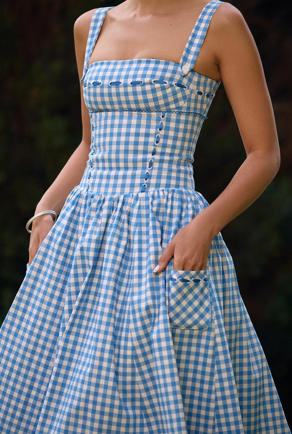 Ariana Sleeveless Midi Sundress in blue-cream gingham with a square neckline, wide straps and a fitted bodice trimmed with ribbon eyelets. Gathered midi skirt with side patch pockets and back zipper; chic and versatile for daytime events.