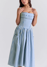 Ariana Sleeveless Midi Sundress in blue-cream gingham with a square neckline, wide straps and a fitted bodice trimmed with ribbon eyelets. Gathered midi skirt with side patch pockets and back zipper; chic and versatile for daytime events.
