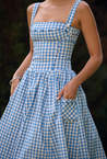 Ariana Sleeveless Midi Sundress in blue-cream gingham with a square neckline, wide straps and a fitted bodice trimmed with ribbon eyelets. Gathered midi skirt with side patch pockets and back zipper; chic and versatile for daytime events.