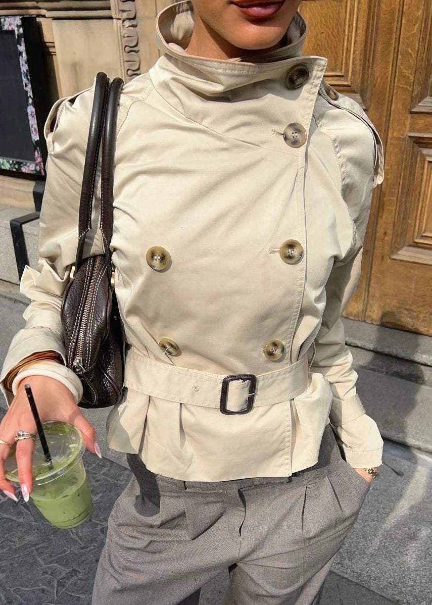 Isabelle high-neck belted trench coat in premium cotton blend with structured stand collar, double-breasted buttons, epaulettes and adjustable cuffs. Tailored silhouette layers over dresses, denim or trousers for refined autumn-winter style.