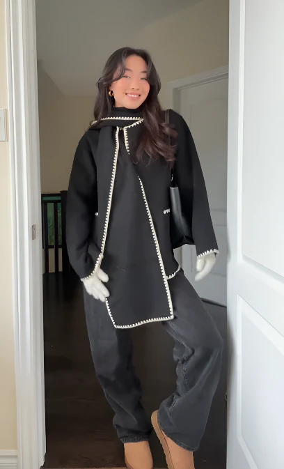 Nellie black wool-blend cape coat featuring a flowing silhouette with white contrast trim, front button fastening, practical side pockets and an attached oversized scarf collar. Warm, elegant outer layer that drapes over denim, knitwear or evening looks.