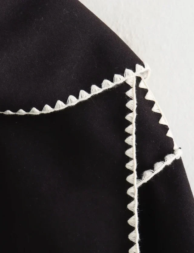 Nellie black wool-blend cape coat featuring a flowing silhouette with white contrast trim, front button fastening, practical side pockets and an attached oversized scarf collar. Warm, elegant outer layer that drapes over denim, knitwear or evening looks.